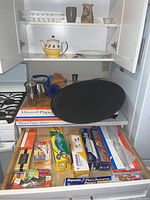 Kitchenware collection including large black oval tray on top of various kitchen wrap boxes, wooden and plastic cutting boards behind, stainless steel cocktail shaker, metal funnel, teapot on shelves above.