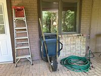 Image showing Werner 6-step aluminum stepladder, single-wheel metal wheelbarrow with black tub, and green garden hose coiled on porch tile floor.