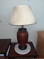 Full view of the lamp showing the pleated off-white shade, reddish-brown wooden base with gold design, and white marble round base.