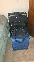 Three luggage pieces: black suitcase, black duffel, blue rolling crate stacked together next to bed.