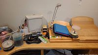 Office supplies on wooden table including calculators, blank CDs in spindles, a white plastic box, desk lamp, wooden angled filing holders, multi-colored folders, and a small box of cables.