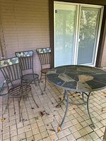 Photo showing the round wrought iron table with stone tile top next to three wrought iron chairs with cushioned backs, placed on a tiled patio.