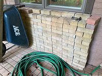 Stack of approximately 200+ light tan paving bricks with a few red bricks on top and side, next to a garden hose and wheelbarrow