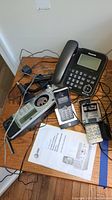 Photo of several corded and cordless phones including handset units and a manual placed on a wooden surface with tangled cords.