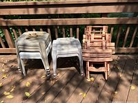 Photo showing three stacks of outdoor plastic tables on a wooden deck; two stacks of white solid plastic tables and one stack of reddish folding frame tables.