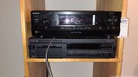 Sony STR-DH100 stereo receiver and Nakamichi CassetteDeck 2 cassette deck stacked on wooden shelf. Receiver has display, master volume knob and control buttons.