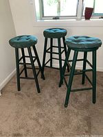 Three green wooden stools with round tufted velvet padded removable seat cushions, shown standing on carpet near a window.