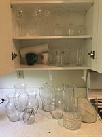 Various glass vases, jar, pitcher on kitchen counter and mugs, paper towels, stemware in cabinets.