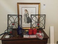 Photo shows framed Cyrano de Bergerac wall art against the wall with two black metal candle holders each containing a red candle, red cylindrical candles in front, a Vietnam-themed mug holding blue pens, scissors, nail clippers, a pocket multi-tool, and a glass heart-shaped container on a wooden surface