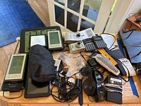 Full overview of lot items on table including clocks, cameras, calculators, sunglasses, and other electronics.