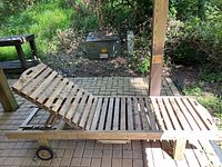 Full side view of the wooden outdoor chaise lounge showing the adjustable slatted backrest, pull-out table underneath, rear wheels, and weathered wood condition.