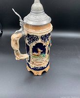 Photo of a detailed ceramic German beer stein with a pewter lid, decorated with a raised relief scene of a deer in a forest and German inscription. The handle resembles carved wood.