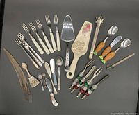 Top-down view of assorted serving utensils including forks, spoons, cake servers, pineapple-shaped handled utensils, and decorative cocktail picks.