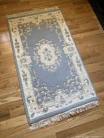 Full view of the rug showing its blue central field with ornate cream border and floral design on hardwood floor.