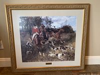 Full view of framed hunting scene with horses and dogs hung on wall.