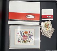 Photo of the Pimpernel branded boxes with 6 square coasters and 8 rectangular placemats featuring floral patterns, arranged on a dark surface.