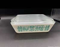 Side view of opaque white Pyrex casserole dish with green farm-themed Butterprint design along side, accompanied by a clear ribbed glass lid.