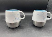 Two white Starbucks coffee mugs with blue snowflake designs and stainless steel bases shown side by side