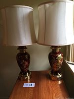 Pair of ceramic table lamps on wooden surface showing Asian-themed design on burgundy bases with fabric shades.