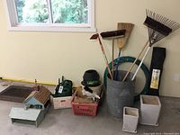 Wide shot showing various garden tools leaning in large metal container, two wooden bird feeders, green watering cans, plastic tray with small tools, metal floor mats, and coiled garden hose.