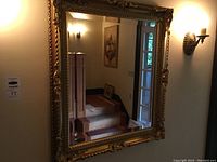 Full view of gold framed mirror hung on wall with reflection of staircase and framed picture.