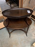 Full view of the two-tier oval side table showing the top removable glass tray with metal handles, the lower wooden shelf, curved legs, and the overall dark wood finish.