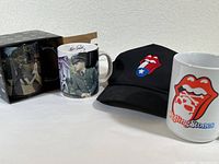 Photo showing one Beatles Abbey Road mug in box, one Elvis Presley mug, one Rolling Stones mug, and a black Rolling Stones hat with logo.