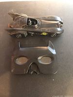 Top view of the black Batmobile car toy and the black Batman mask showing their shapes and logos.