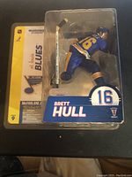 Photo displaying the Brett Hull McFarlane figure in packaging, showing the figure dressed in St. Louis Blues blue jersey with number 16 and helmet. Packaging includes NHL and McFarlane branding visible on the backing card.