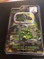 Front of original package showing Incredible Hulk digital camera with three interchangeable faceplates in blister pack. Displays green and black Hulk graphics with yellow accents. Slight package damage visible.