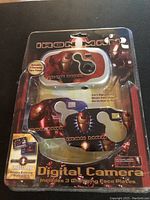 Front view of the Iron Man digital camera in original packaging showing the white and red camera body with three interchangeable face plates featuring Iron Man helmet designs.