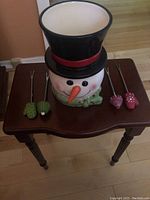 Snowman fondue set assembled on wooden table showing snowman base with black top hat lid and four fondue forks with mitten handles.
