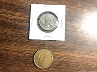 Two American coins on a wooden surface; the 1976 bicentennial quarter in a protective cardboard holder and the Sacagawea dollar coin loose.