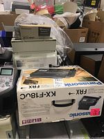 Photo showing Panasonic KX-F180C fax machine box with Brother Intellifax 1270 fax machine next to it, partially covered items around.