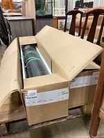 Open cardboard boxes on a pallet containing two black flooring rolls in protective foam