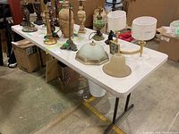 White folding plastic table with metal legs supporting various lamps and lamp parts, visible scratches and some stains on table surface.