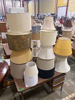 Photo showing assorted lampshades stacked and arranged on wooden furniture in a warehouse setting.