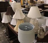 View of lamp shades arranged on wooden dresser, showing variety of medium to large fabric shades including white, cream, and brown colors plus smaller wrapped shades.
