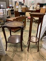 Both wooden plant stand and side table shown in a warehouse setting with lamps and other furniture.