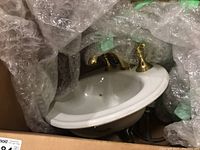 Photo showing one white porcelain Kohler sink with gold tone brass faucet, partially wrapped in bubble wrap inside box.