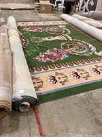 Partial unrolled view showing full width of green floral pattern rug and other rolled rugs surrounding it.