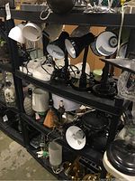 Photo showing the utility shelving loaded with various lamps and light fixtures for context. Shelving is black plastic with three shelves and round legs.