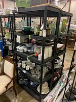 Four-tier black plastic shelving unit loaded with various glassware and dishware in a warehouse setting.