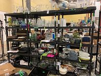 Three black plastic shelving units filled with various glassware and plates in a warehouse setting.