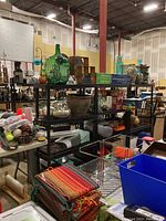 Three black plastic shelving units holding various items like pottery, rugs, boxes, and miscellaneous storage items within a large indoor space.