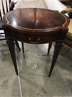 Front view of the wood side table showing scalloped top with inlay and front drawer with brass handle.