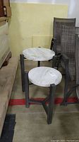 Photo showing two round side tables with faux marble composite tops and dark angled faux wood legs in a warehouse setting.