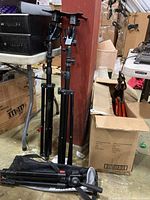 Two black Mantis support stands and folded Manfrotto camera tripod standing on a floor near boxes and tables.