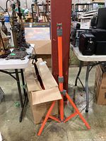 Single assembled orange work light stand displayed upright next to its packaging box on warehouse floor.