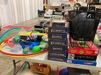 Table with multiple board games in boxes stacked, assorted hula hoops, toy guns, black basket, and puzzle box visible.